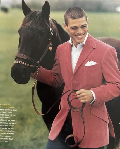 Man in a pink corduroy jacket holding a horse, smiling outdoors.