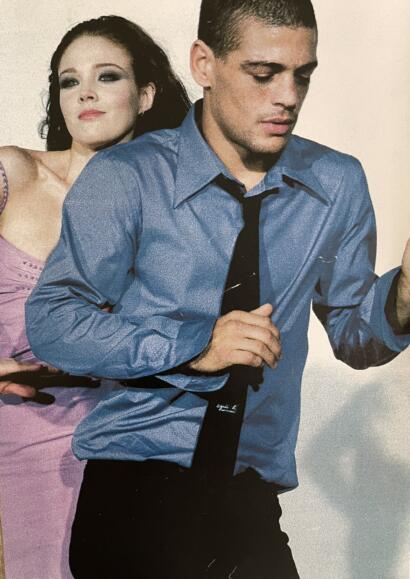 Stylish young man in a blue shirt and black tie, with a woman in a pink dress in the background.