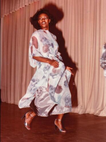 Model in metallic floral gown with heels, dancing on stage with a beige curtain backdrop.