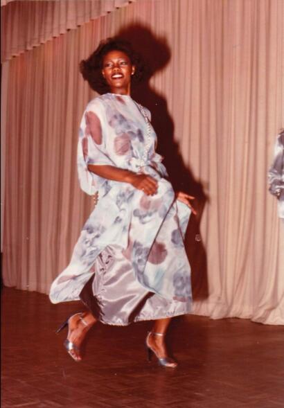 Model in metallic floral gown with heels, dancing on stage with a beige curtain backdrop.