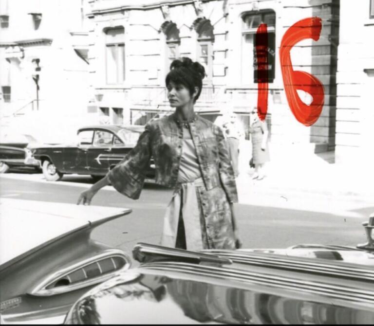 Stylish woman in a vintage city street, dressed in a patterned jacket and high-waisted skirt, with a bold red "16" painted on the wall behind her.