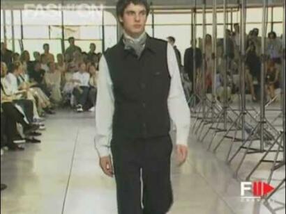 A model walks down an indoor runway in a black vest and white shirt, with an audience in the background.
