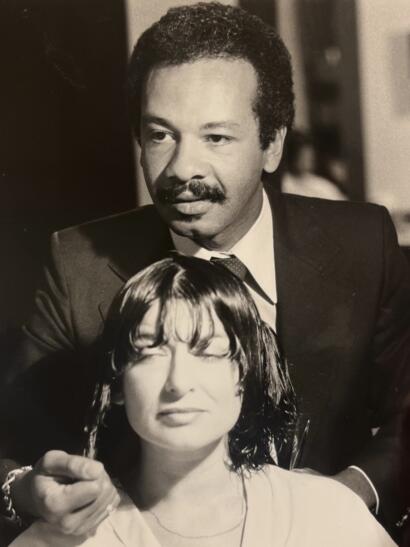 Black-and-white photo of a man with a mustache in a suit standing behind a woman with wet hair and closed eyes.