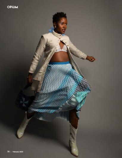 Model in a layered outfit with a pleated blue patterned skirt, cream coat, white boots, and accessories.