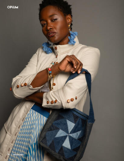 Elegant woman in beige jacket with colorful buttons, accessorized with blue earrings and bracelets, holding a geometric tote bag.