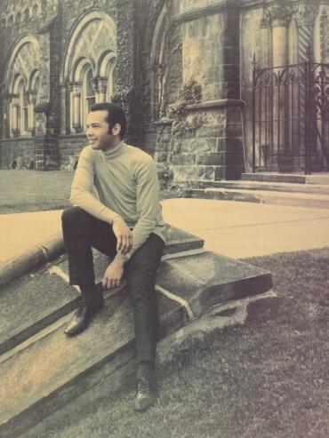 A man in a light turtleneck and dark pants seated on a stone bench outside a historic building, styled in a vintage black-and-white photo.