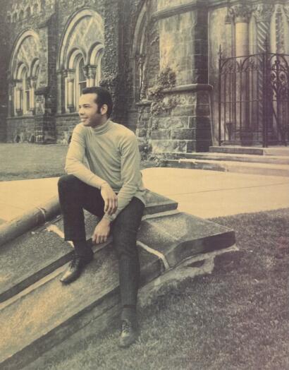 A man in a light turtleneck and dark pants seated on a stone bench outside a historic building, styled in a vintage black-and-white photo.