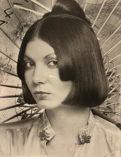 A woman with sleek, straight bob haircut and traditional embroidered blouse, set against a decorative parasol.