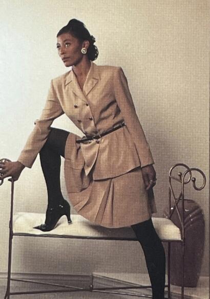 Woman in a beige business suit with navy tights, black high heels, and bold earrings.