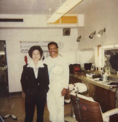 A man and a woman stand together in a room, both smiling, with the woman wearing a black outfit with white accents and the man dressed in a light-colored outfit.