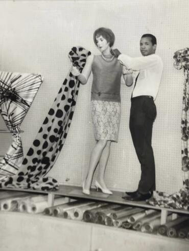 A mannequin displays a patterned dress with a pearl necklace, assisted by a man in a white shirt.