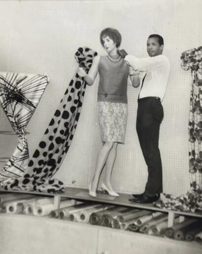 A mannequin displays a patterned dress with a pearl necklace, assisted by a man in a white shirt.