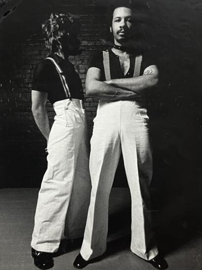 Two women in high-waisted wide-leg pants and short-sleeve tops pose against a brick wall.