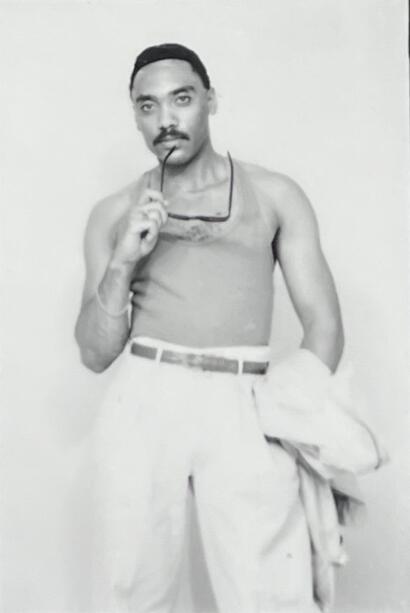 Man with sunglasses in hand, wearing a sleeveless shirt and trousers, standing against a plain background.