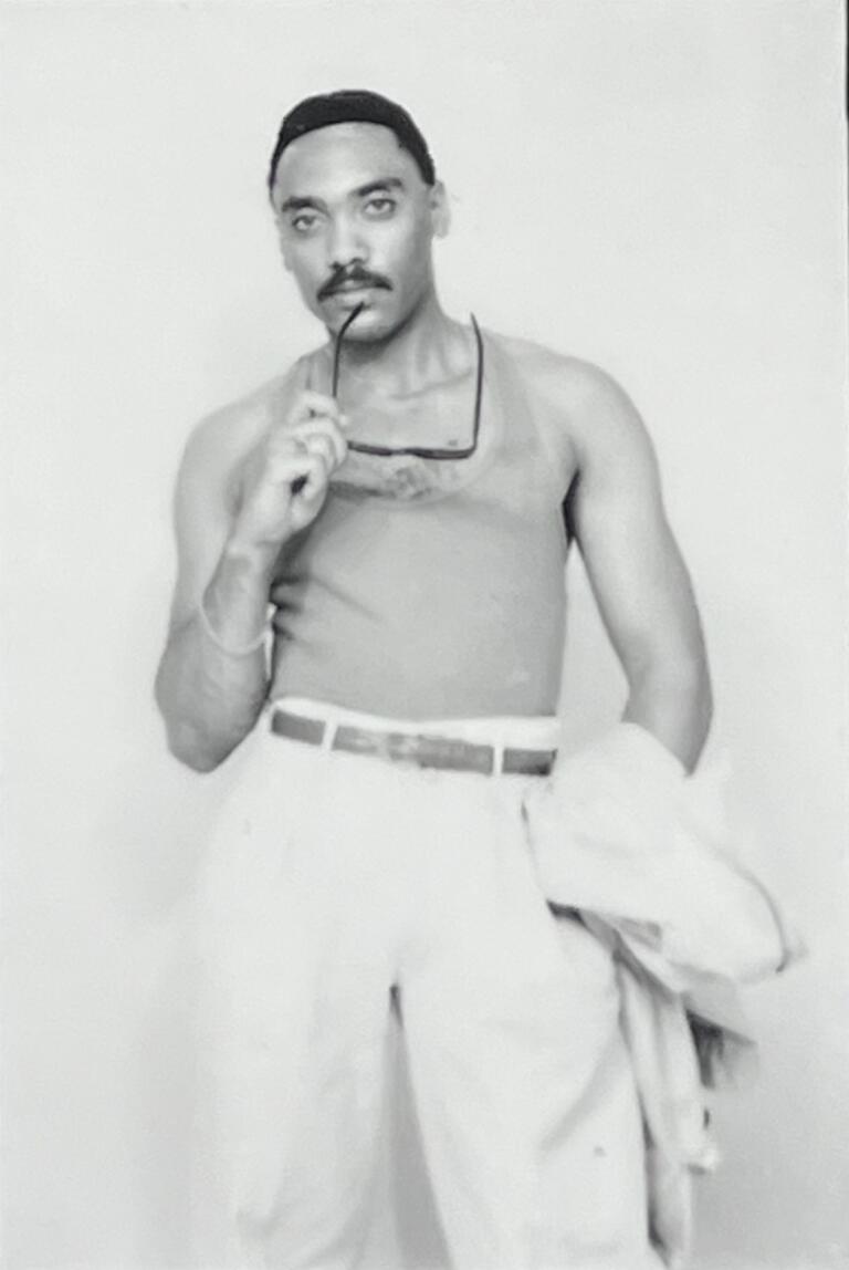 Man with sunglasses in hand, wearing a sleeveless shirt and trousers, standing against a plain background.