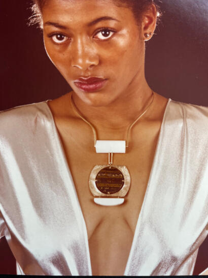 Close-up of a woman with natural makeup, wearing a striking statement necklace and a shiny, satin-like silver top.