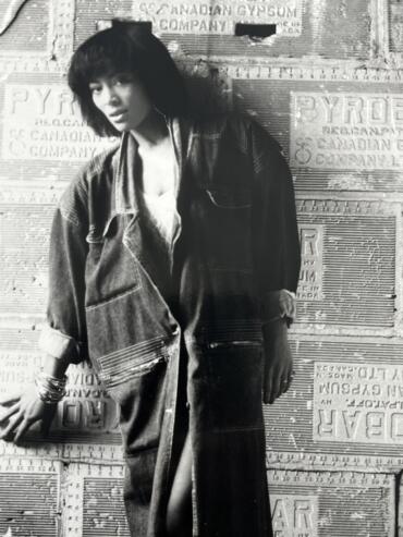 A black-and-white portrait of a woman in casual denim, standing against a textured, vintage advertisement background.