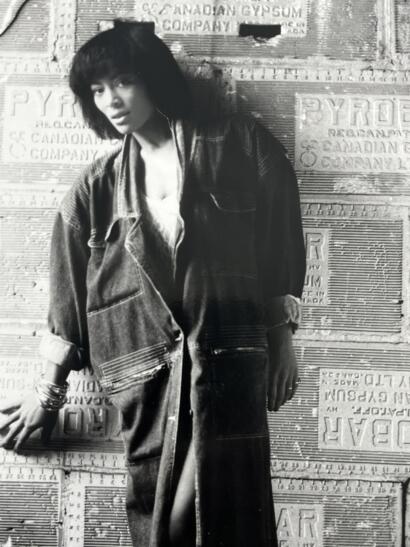 A black-and-white portrait of a woman in casual denim, standing against a textured, vintage advertisement background.