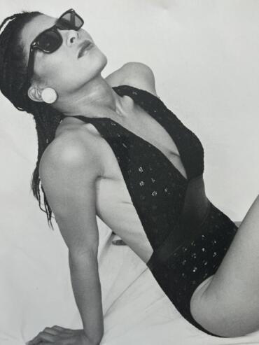 Black and white image of a woman in sunglasses, wearing a lace top and large earrings, posing confidently.