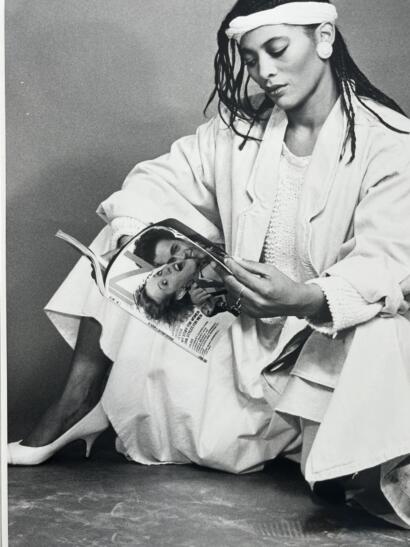 Black-and-white portrait of a woman reading a magazine, wearing a light-colored coat, high heels, and large earrings.