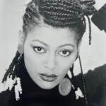 Black and white portrait of a woman with styled dreadlocks, wearing earrings and a buttoned-neck top.
