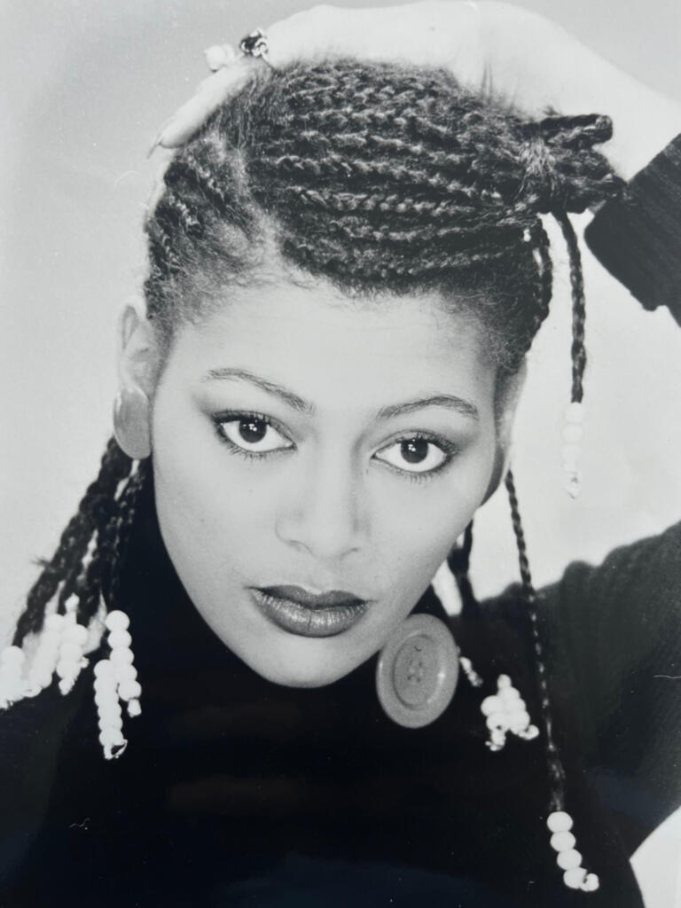 Black and white portrait of a woman with styled dreadlocks, wearing earrings and a buttoned-neck top.