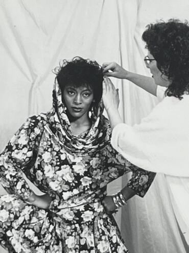 Model with short curly hair and bold makeup being styled by a stylist in a floral outfit.