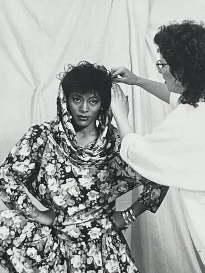 Model with short curly hair and bold makeup being styled by a stylist in a floral outfit.