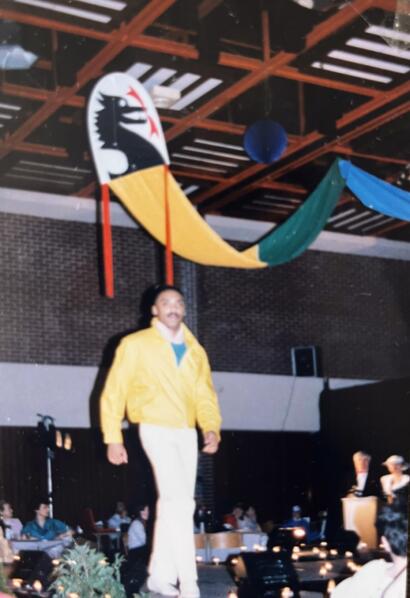 Man in a yellow jacket standing on a stage with colorful drapes and decorations overhead.