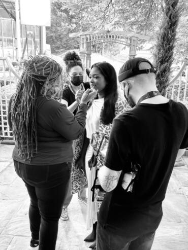 Model getting makeup touch-up outdoors, surrounded by team members.