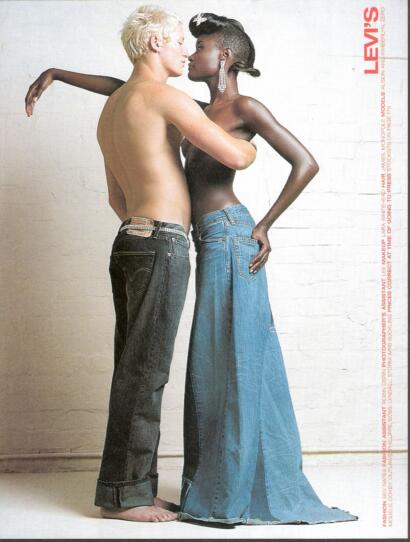 Two models, one with platinum hair and the other with dark skin and styled hair, pose close together against a white brick wall, wearing denim jeans.