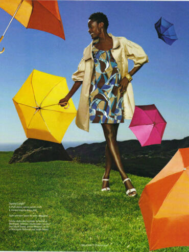 Woman in a colorful patterned dress and beige puff-sleeve jacket holding a yellow umbrella, surrounded by other colorful umbrellas under a blue sky.