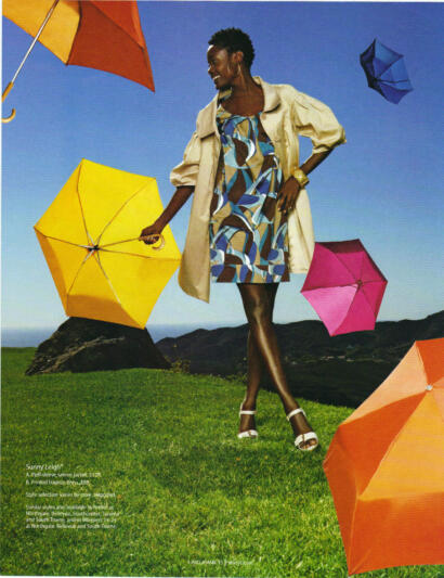 Woman in a colorful patterned dress and beige puff-sleeve jacket holding a yellow umbrella, surrounded by other colorful umbrellas under a blue sky.