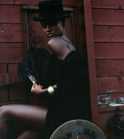A woman in a black hat and sheer dress holding a black fan, against a rustic wooden background.