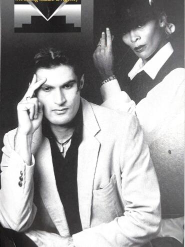 Black and white promotional photo of two fashion models, one male and one female, posing with a "Visual Impact" modeling studio sign in the background.