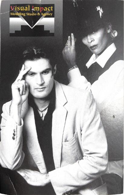 Black and white promotional photo of two fashion models, one male and one female, posing with a "Visual Impact" modeling studio sign in the background.