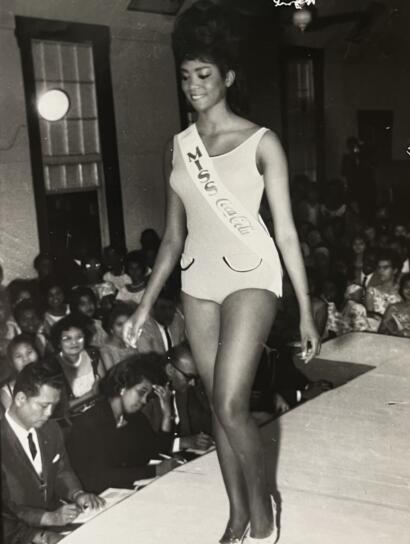 Vintage black-and-white photo of a Miss Coca-Cola beauty contestant walking on stage, wearing a sash and a fitted, sleeveless dress.