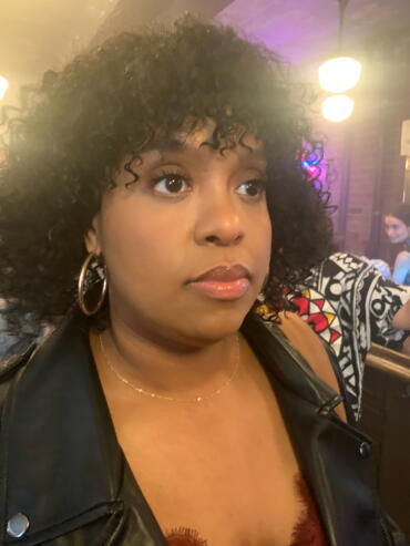 Close-up of woman with curly hair, hoop earrings, and a black leather jacket in a dimly lit setting.