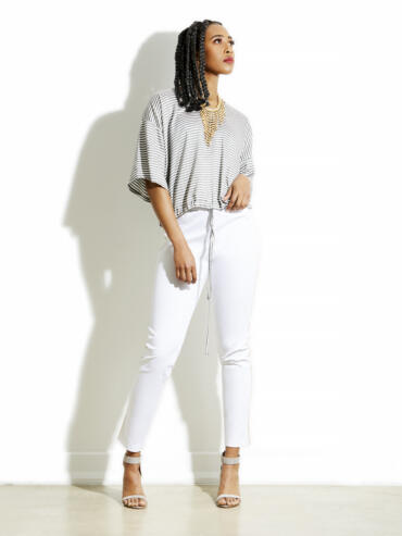 Woman in a striped top and white pants, wearing heels and a statement necklace, standing against a white wall.