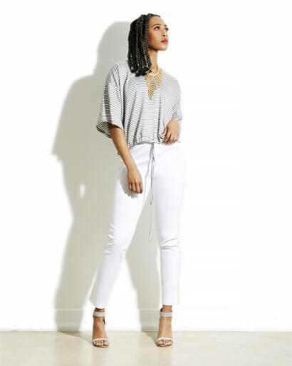 Woman in a striped top and white pants, wearing heels and a statement necklace, standing against a white wall.