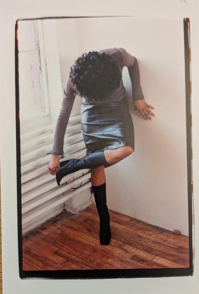 A person in a black satin skirt, knee-high boots, and a long-sleeve top, posed against a white wall with wooden flooring.