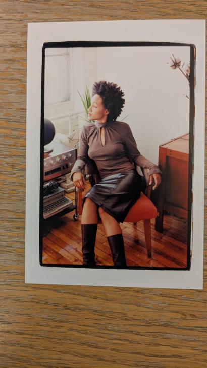A woman with natural hair sits gracefully in a cozy studio, dressed in a chic brown top and leather skirt.
