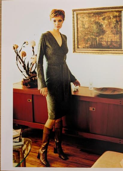 Woman in a gray knit dress and tall boots standing beside a wooden sideboard in an art-filled room.