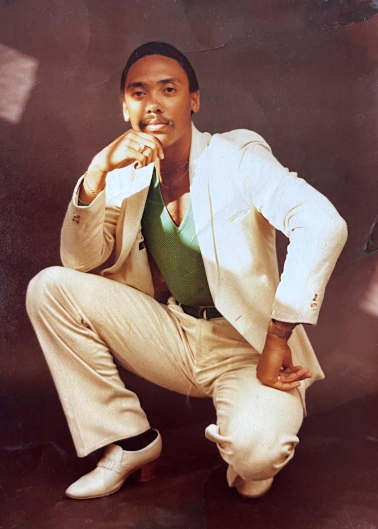 Man in an elegant cream suit with a green shirt, seated in a confident pose.