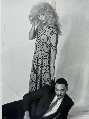 Black-and-white photo of a woman with voluminous curly hair in a patterned dress standing, and a man in a suit reclining on the floor.