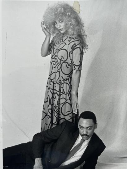 Black-and-white photo of a woman with voluminous curly hair in a patterned dress standing, and a man in a suit reclining on the floor.