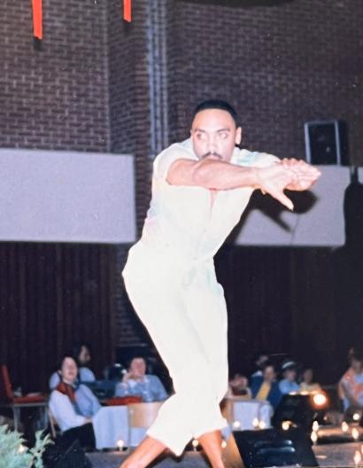 A performer in a white outfit strikes a pose on stage, with an audience seated behind.