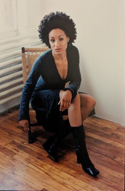Woman seated on a wicker chair in a stylish black dress with knee-high boots.