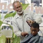 Jimmy Wisdom stands inside the storefront window of a barbershop, taking the clippers to the hair of a young boy.