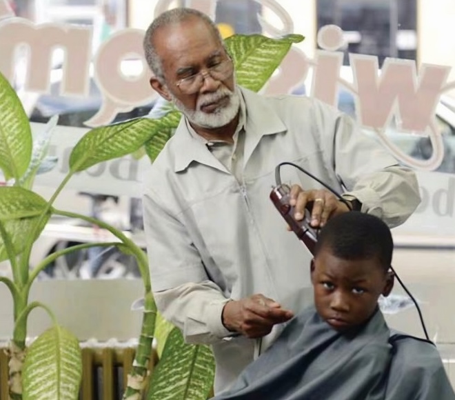 Jimmy Wisdom stands inside the storefront window of a barbershop, taking the clippers to the hair of a young boy.